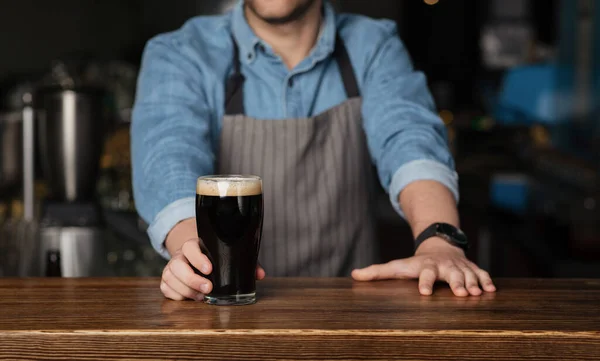 Barman en camisa de mezclilla y delantal está detrás de un mostrador de madera y le da un vaso de cerveza oscura — Foto de Stock