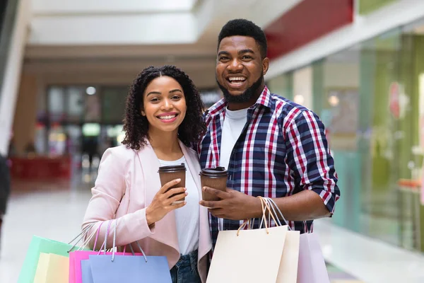 Feliz pareja negra posando en el centro comercial con bolsas de compras y café —  Fotos de Stock