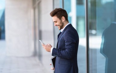 Gülümseyen CEO, büyük şehirde gökdelenin yakınında, cep telefonuyla ortağıyla iletişim kuruyor.