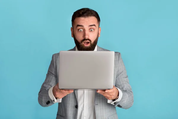 Lottogewinner. Jungunternehmer blickt überrascht auf Laptop-Bildschirm auf blauem Hintergrund — Stockfoto