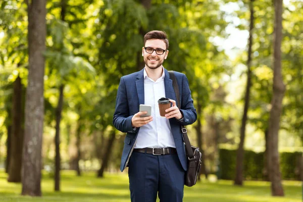 Happy office worker with his morning coffee checking emails on phone at park on sunny day — Stock Photo, Image