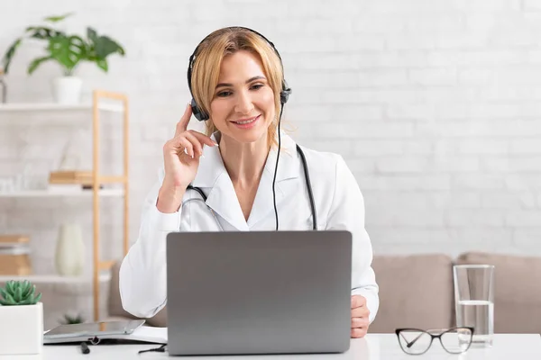 İnceleme ve tanı çevrimiçi. Gülümseyen doktor kulaklığa dokunur ve laptopa bakar. — Stok fotoğraf