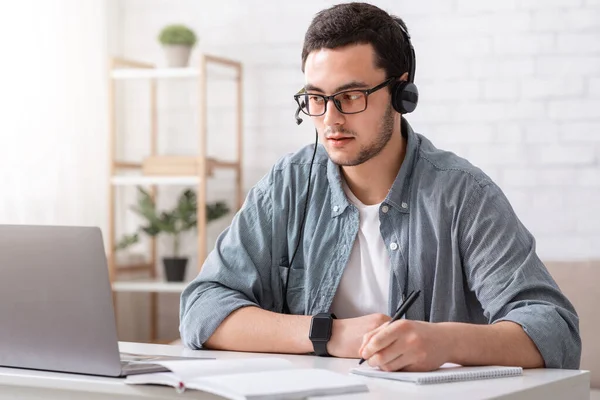 Professora online. Cara de óculos e fone de ouvido faz anotações no notebook e olha para o laptop — Fotografia de Stock