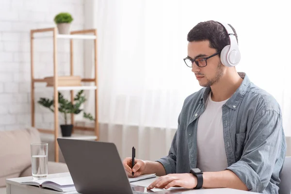 Elfoglalt fickó szemüveggel és fejhallgatóval jegyzetel a jegyzetfüzetbe, és laptopon dolgozik a nappaliban. — Stock Fotó