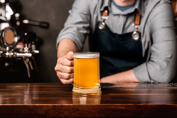 Cantinero amistoso. Hombre en delantal pone una taza de cerveza ligera en el mostrador de madera en el interior — Foto de Stock