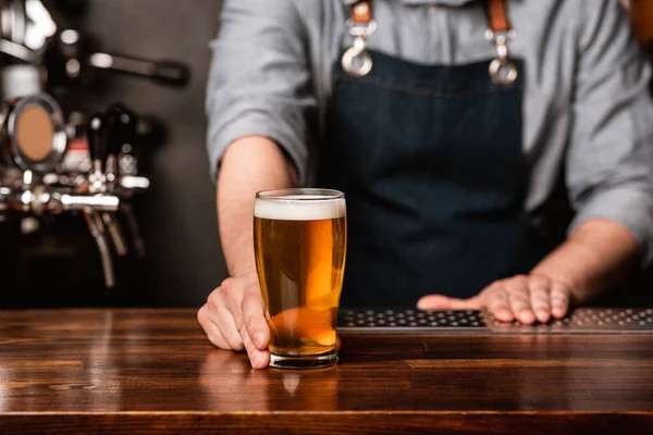 Uitgifte van bier aan de bar. Barman in schort geeft de klant een glas licht bier op houten toonbank — Stockfoto