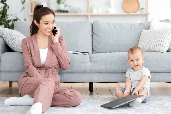 Cheerful Mom Talking On Cellphone While Baby Playing With Laptop At Home