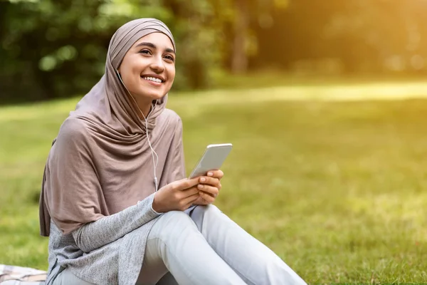 Chica árabe alegre en hijab escuchando música en el parque —  Fotos de Stock