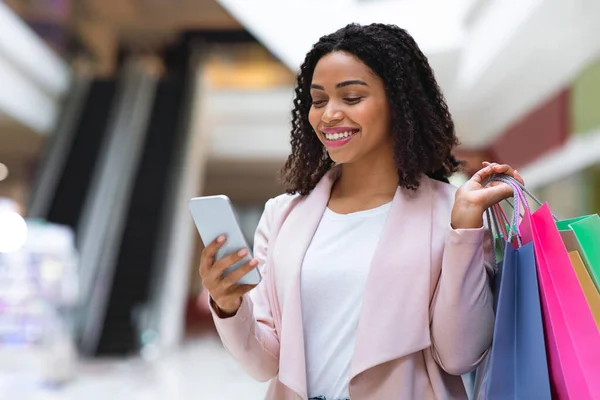 割引アプリ。ショッピングバッグで笑顔黒女性ショッピングバッグ使用スマートフォンでモール — ストック写真