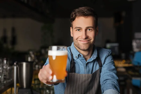 Pinta cerveza. Barman sonriente en delantal sostiene un vaso de cerveza ligera en el interior del pub — Foto de Stock