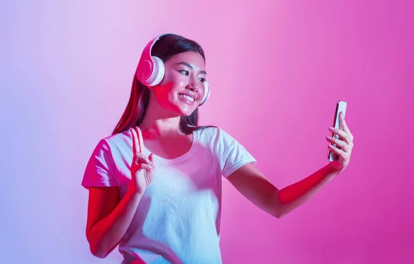 Music and selfie. Smiling girl in headphones shows peace sign and makes photo — Stock Photo, Image