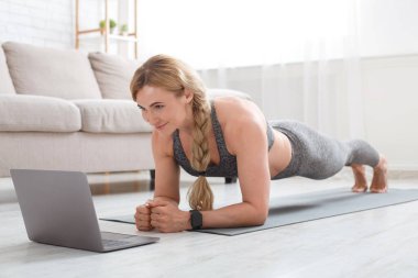 Fitness egzersizi. Spor giysili bir kadın paspasa tahta örüyor ve dizüstü bilgisayara bakıyor.