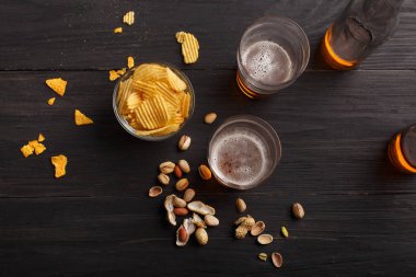 Siyah masadaki cam kasede çipler ve şam fıstıkları, yanında hafif bira ve şişe olan iki bardak.