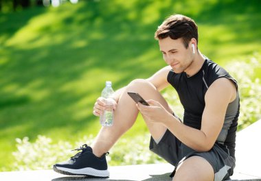 Kablosuz kulaklıklı adam akıllı telefona bakıyor, su dolu bir şişe tutuyor, parkta oturuyor.