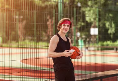 Dışarıda basketbol sahasında cep telefonu ve topu olan neşeli bir milenyum sporcusu.
