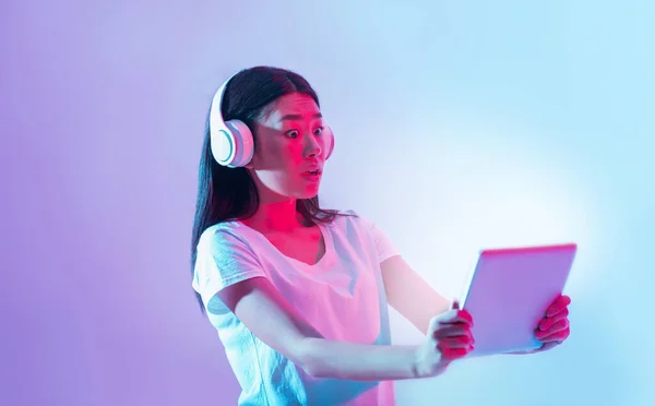 Les appareils modernes sont surpris. Choqué asiatique fille en blanc casque regarde tablette avec écran lumineux — Photo