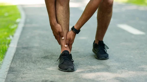 Sports injury. Closeup of millennial guy holding his hurt ankle on jogging track at park — Stock Photo, Image