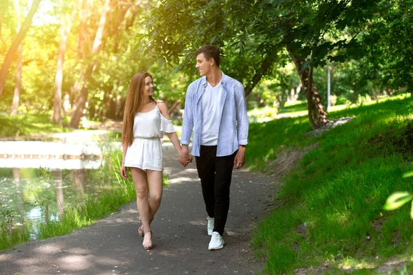 Ласковая пара тысячелетия, держащаяся за руки и идущая по дорожке в летнем парке — стоковое фото