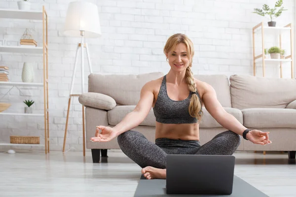 Online fitness lessons. Middle aged woman practicing home yoga with laptop in living room — Stock Photo, Image