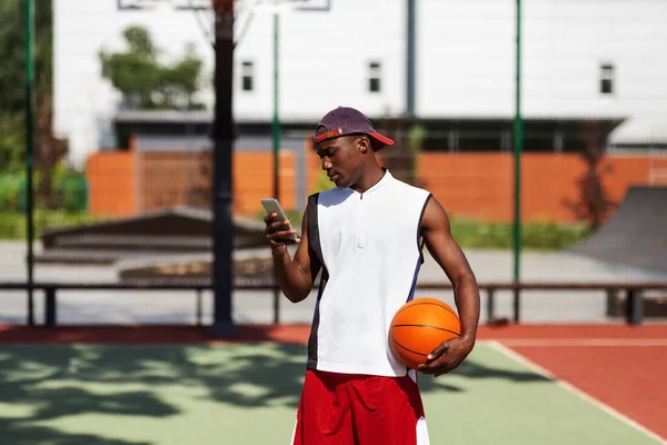 Svart idrottsman surfa på nätet på mobiltelefon innan basketmatch på utomhus arena — Stockfoto