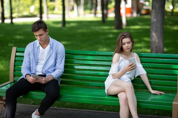 Gadget adicción y problemas de relación. Pareja joven con teléfonos inteligentes sentados en el banco en el parque, ignorándose mutuamente —  Fotos de Stock