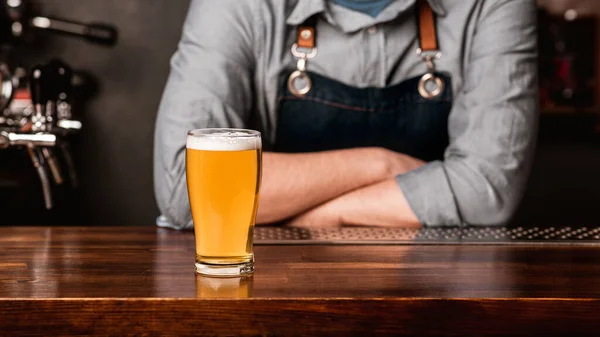 Habla con el camarero con bebida. Hombre en delantal con brazos cruzados, y cerveza ligera con espuma en barra de madera — Foto de Stock