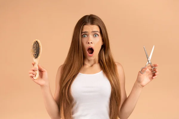 Surpreendida menina milenar com cabelos longos e lisos segurando tesoura e escova no fundo bege — Fotografia de Stock