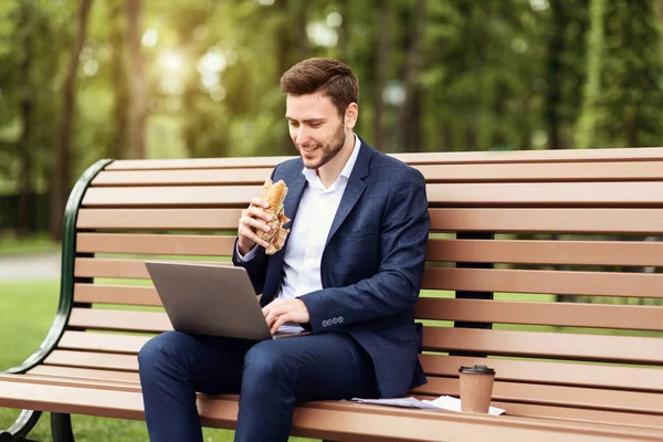 Millennial businessman eating sandwich and working on laptop during lunch break at park — Stock Photo, Image