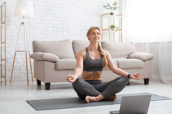 Mujer en ropa deportiva en posición de loto meditación en la estera en el interior de la sala de estar delante de la computadora portátil —  Fotos de Stock