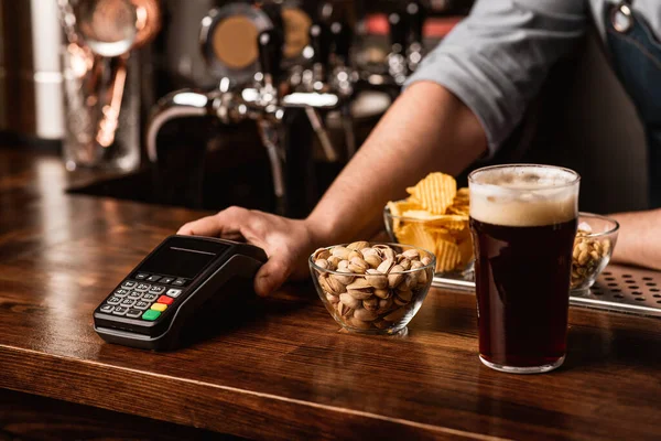 Barman geeft contactloze terminal, in de buurt pistaches, chips en noten in borden, en donker bier met schuim — Stockfoto