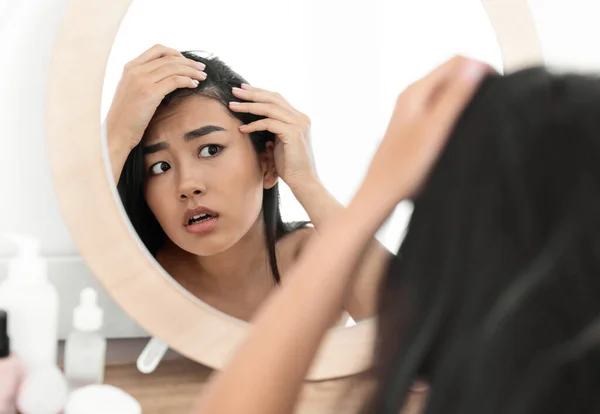 Ανήσυχη Ασιάτισσα που κοιτάει τα μαλλιά της στον καθρέφτη στο σπίτι. — Φωτογραφία Αρχείου