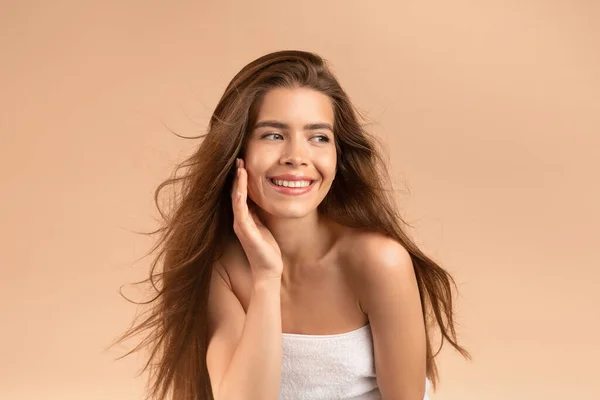 Sexy giovane signora con pelle pura perfetta e capelli castani setosi sorridente e toccando il suo viso su sfondo beige — Foto Stock