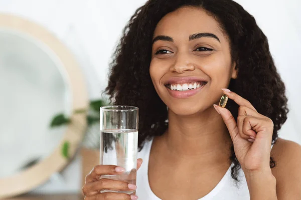 Ingesta diaria de vitaminas. Mujer negra feliz sosteniendo la píldora de la cápsula y el vaso de agua —  Fotos de Stock