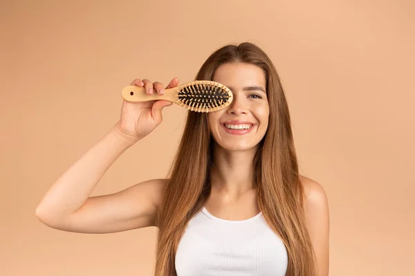 Retrato de senhora bonita com cabelos longos e sedosos e escova de madeira sorrindo no fundo bege — Fotografia de Stock