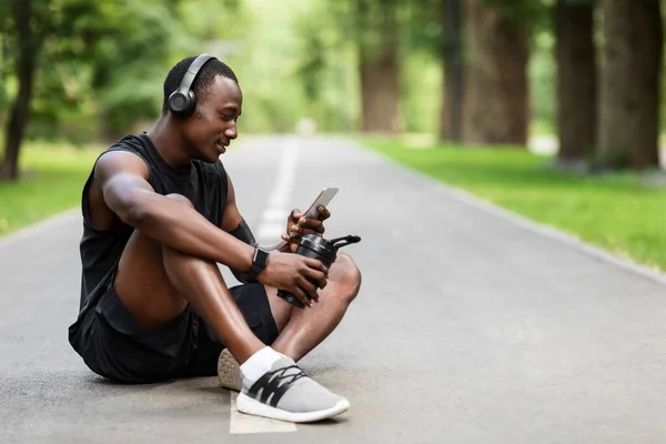 Park yolunda oturan, içki içen ve telefon kullanan yorgun sporcu. — Stok fotoğraf