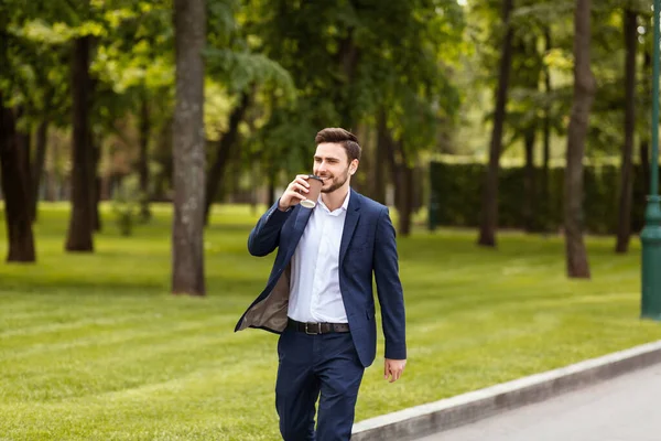 Smiling entrepreneur drinking his morning coffee from paper cup during walk in park — Stock Photo, Image