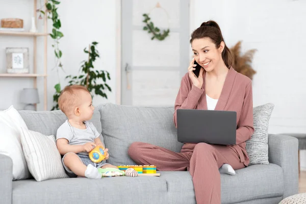 Mujer hablando en el teléfono celular y el uso de la computadora portátil mientras cuida de su hijo pequeño — Foto de Stock