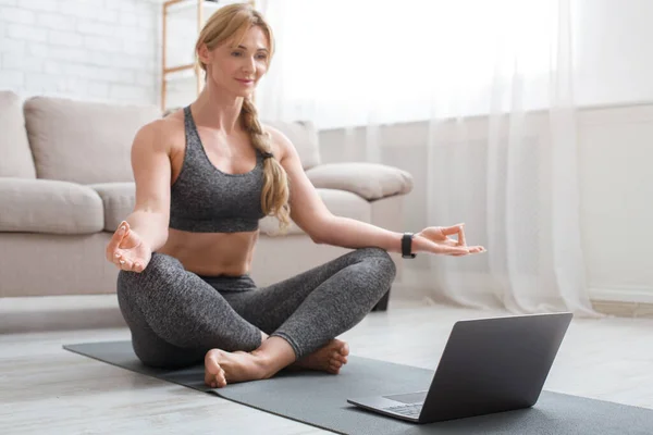Fitness, yoga y pilates. Mujer de mediana edad en ropa deportiva haciendo yoga con portátil en la estera —  Fotos de Stock