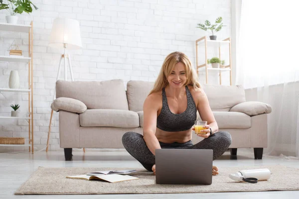 Lächelnde Frau mit einem Glas Saft, benutzt Laptop, sitzt mit Notizblock und Flasche im Wohnzimmer auf dem Boden — Stockfoto
