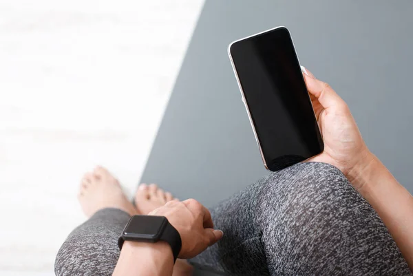Mujer en pantalones de chándal sentada en el suelo en la estera con reloj inteligente, sosteniendo el teléfono inteligente con pantalla en blanco en la mano — Foto de Stock