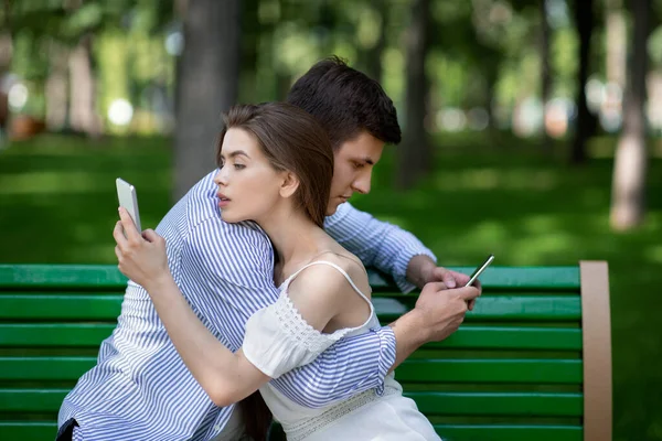 Gadget závislý pár pomocí mobilních telefonů při objímání na lavičce v parku — Stock fotografie