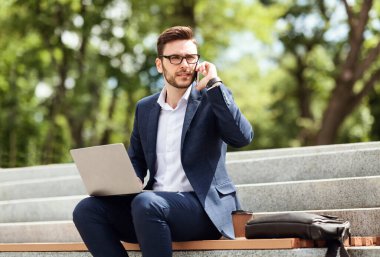 Genç CEO bilgisayarda dokümanları karıştırıyor ve parktaki merdivenlerde telefon görüşmesi yapıyor.