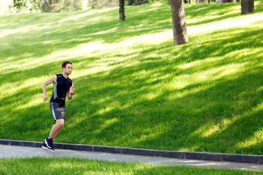Genç adam güneşli bir günde koşar. Yeşil çimlerle parkta koşan sporcu