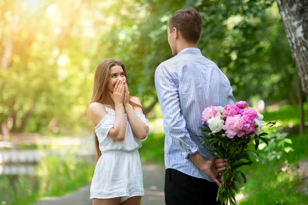 Tierno novio escondiendo ramo de hermosas peonías para su chica a sus espaldas en el parque — Foto de Stock