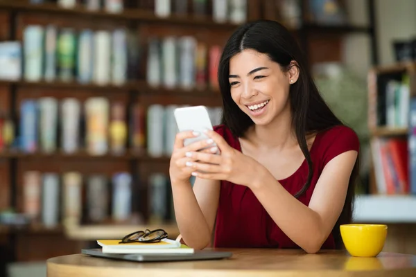 Communication en ligne. Heureuse fille asiatique en utilisant smartphone tout en passant du temps au café — Photo