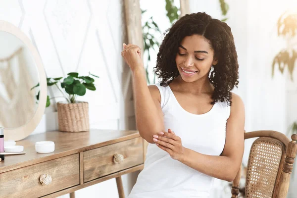 Tratamiento de codos secos. Sonriente mujer negra aplicando crema hidratante en casa — Foto de Stock