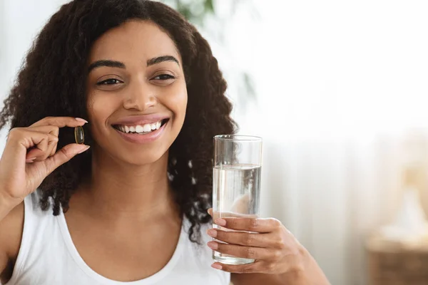 Concepto de bienestar. Sonriente mujer africana sosteniendo cápsula multivitamínica y vaso de agua —  Fotos de Stock