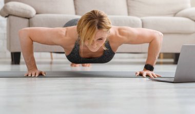 Evde şınav çek. Yetişkin bir kadın laptopla yerde spor yapıyor.