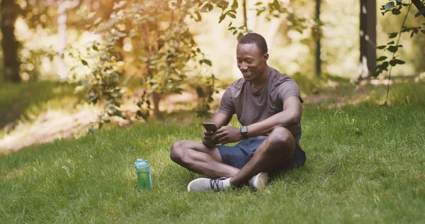 Chico afroamericano usando teléfono inteligente, sentado en la hierba en el parque — Foto de Stock