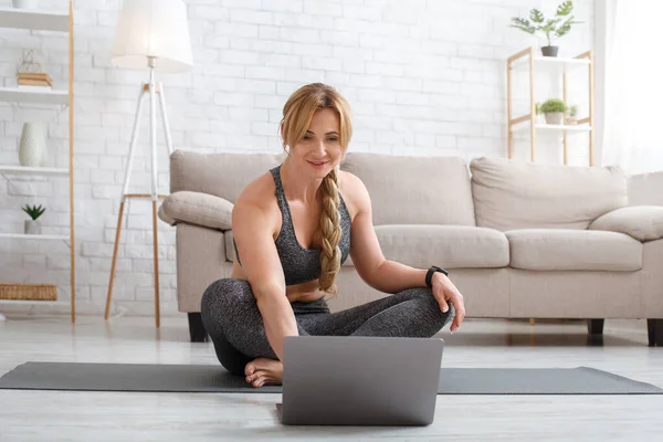 Online stream workout. Woman sitting in lotus pose with laptop in living room interior